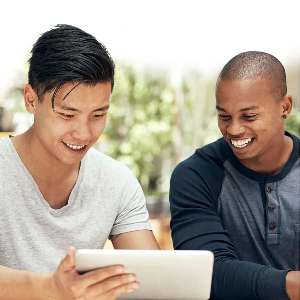 Two friends smiling while using a tablet outdoors.